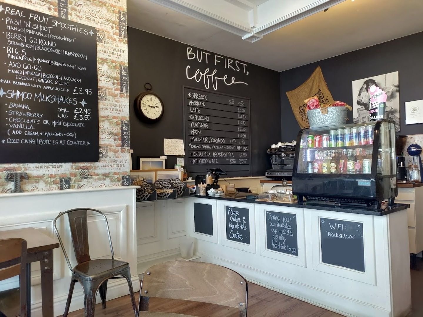 Inside Bradshaw’s: a white panelled counter with small chalkboards, a drinks fridge, an espresso machine, a dark wall chalked ‘But first, coffee’ and a smoothie board on brick-print wallpaper