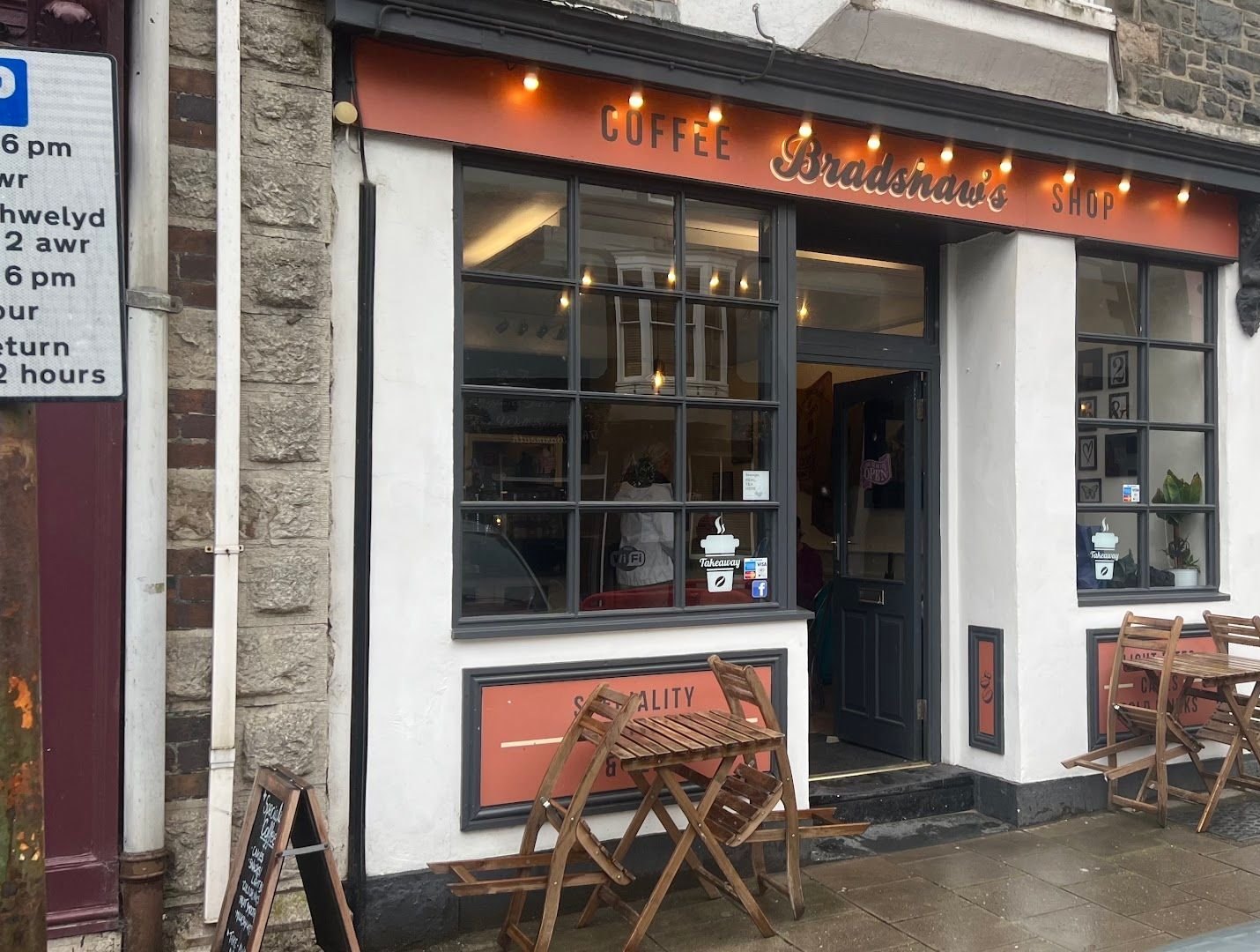 Bradshaw’s from the street: an orange fascia with a string of lights, big windows, and wooden bistro tables and chairs on the pavement